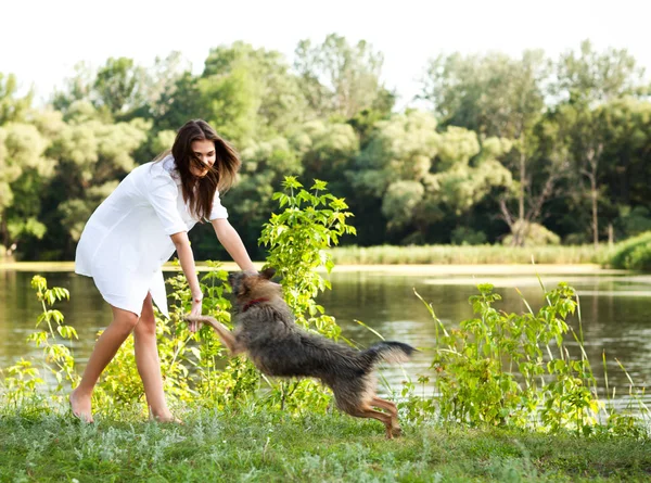 Junge brünette Frau im weißen Kleid spielt mit Hund auf Gras an einem Sommertag mit grünen Bäumen und Fluss im Hintergrund — Stockfoto