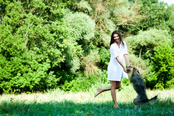 Молодая брюнетка в белом платье играет с собакой на траве в летний день с зелеными деревьями и рекой на заднем плане — стоковое фото