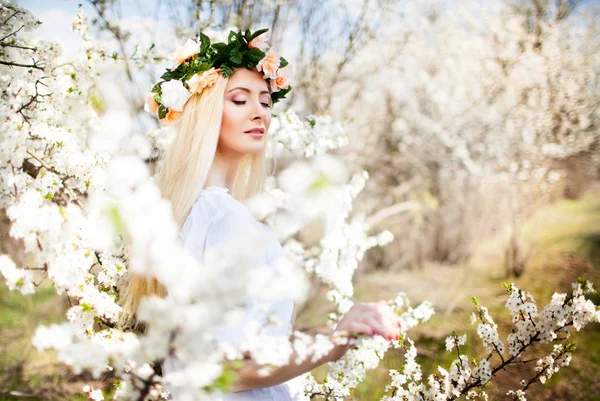 Genç, güzel, sarışın, gülümseyen, beyaz elbiseli bir kadın. Çiçek açan ağaçlarla arka planda duruyor. — Stok fotoğraf