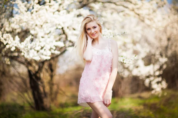 Ung vacker blond leende kvinna i vit mini klänning står med blommande träd i bakgrunden — Stockfoto