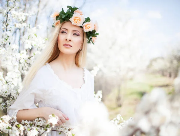 年轻美丽的金发女郎，身穿白衣，头戴玫瑰花环，站在郁郁葱葱的树上，望着旁边 — 图库照片