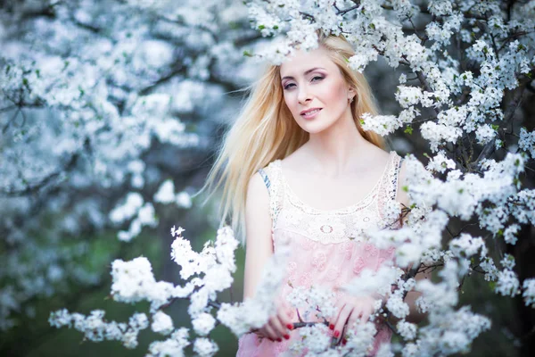 Young beautiful blonde smiling woman in white mini dress standing with blooming trees at background — 스톡 사진