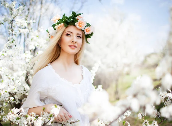 Jonge mooie blonde glimlachende vrouw in witte jurk en roos krans staan in bloeiende bomen en kijken opzij — Stockfoto