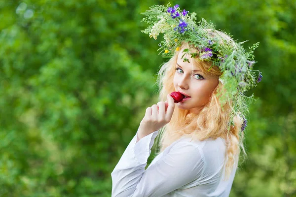 Young beautiful blond woman in white dress and floral wreath standing and eating strawberry on summer day — Stock Photo, Image