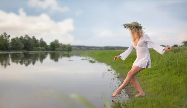 Young sexy beautiful blond woman in white mini dress and floral wreath standing and trying water on summer day — 스톡 사진