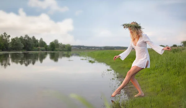 Young sexy beautiful blond woman in white mini dress and floral wreath standing and trying water on summer day — Stock Photo, Image