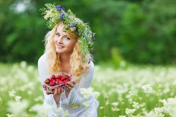 Young beautiful blond woman in white dress and floral wreath holding box of fresh strawberries on summer day — Stock Photo, Image