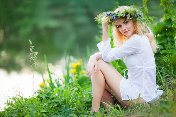 Young beautiful blond woman in white mini dress and floral wreath sitting near lake and smiling on summer day — Stock Photo, Image