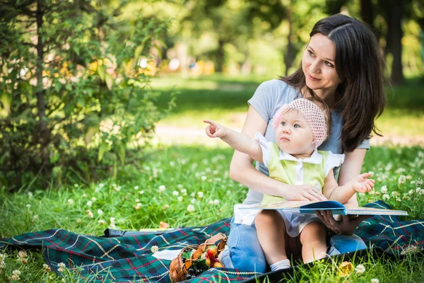 Mãe sentada na grama com sua filha pequena e olhando para o livro — Fotografia de Stock