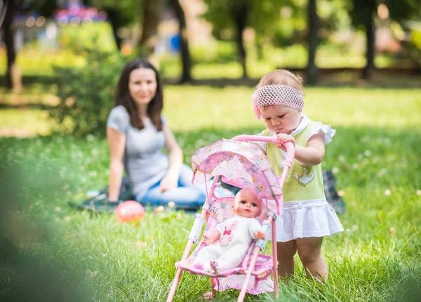 Mor sitter på gräs och hjälpa dotter att gå och rulla leksak barnvagn — Stockfoto