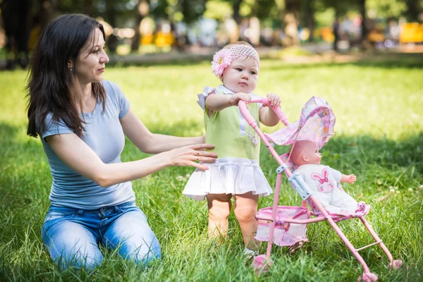 Mor sitter på gräs och hjälpa dotter att gå och rulla leksak barnvagn — Stockfoto