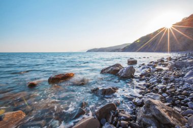 Güneşli bir yaz sabahında, yüksek dağların ve tepelerin arka planında dalgaların kayalık bir sahil boyunca sıçradığı güzel bir deniz manzarası. Seyahat konsepti. Reklam alanı