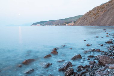 Güneşli bir yaz sabahında, yüksek dağların ve tepelerin arka planında dalgaların kayalık bir sahil boyunca sıçradığı güzel bir deniz manzarası. Seyahat konsepti. Reklam alanı
