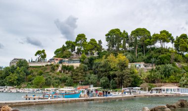 Korfu Adası, Yunanistan - 4 Ekim 2019: Korfu adasında tekneleri olan deniz kenarı, Yunanistan. Turistler için ağaçlar ve oteller olan İyon deniz plajının güzel manzarası, iskelede yürüyen insanlar..
