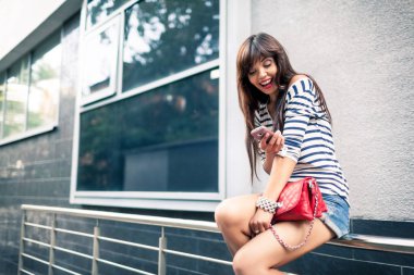 Dışarıda cep telefonu kullanan uzun saçlı güzel bayanın yan görüntüsü. Çizgili tişörtlü çekici kız akıllı telefondan mesajları kontrol ediyor. Mobil iletişim kavramı.