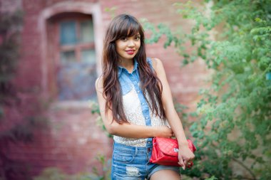 Önde, uzun saçlı, kameraya bakıp gülümseyen güzel bir genç kadın var. Kot şortlu hoş bayan elinde zarif kırmızı bir çanta tutuyor. Güzellik ve tarz kavramı.