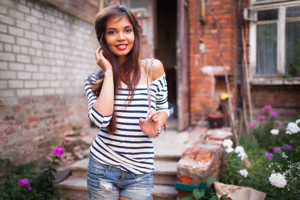 Front view of attractive joyful lady touching hair and looking at camera with smile. Charming young woman wearing stylish striped shirt and denim shorts. Concept of beauty and style.