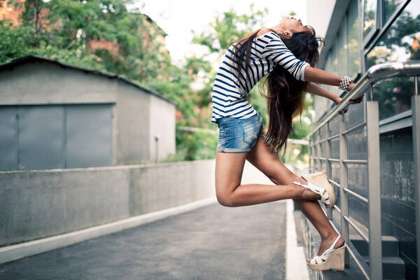 Side view of beautiful young lady gripping handrails and bending backwards on the street. Pretty girl with long hair wearing striped shirt and denim shorts. Concept of freedom.
