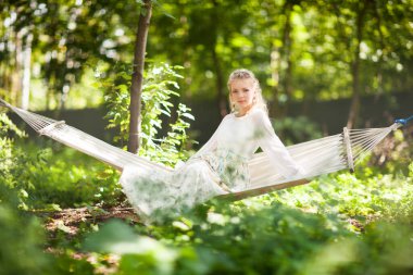 Bahar ormanında hamak salıncağında dinlenen güzel genç bir kadının ön görüntüsü. Zarif, uzun elbiseli, kameraya bakan hoş bir bayan. Güzellik, zarafet ve doğa kavramı.