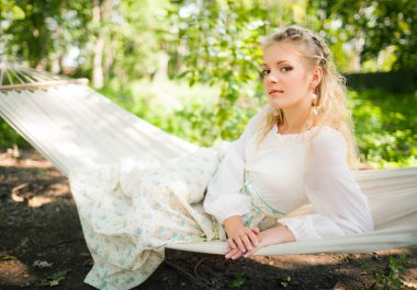Bahar ormanında hamak salıncağında dinlenen güzel genç bir kadının ön görüntüsü. Zarif, uzun elbiseli, kameraya bakan hoş bir bayan. Güzellik, zarafet ve doğa kavramı.
