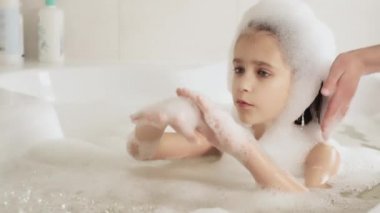 Küçük kız küvette banyo yapıyor ve köpükle oynuyor, hijyen ve bebek bakımı konsepti, 4K UHD hala video kamera, 2x yavaş çekim.