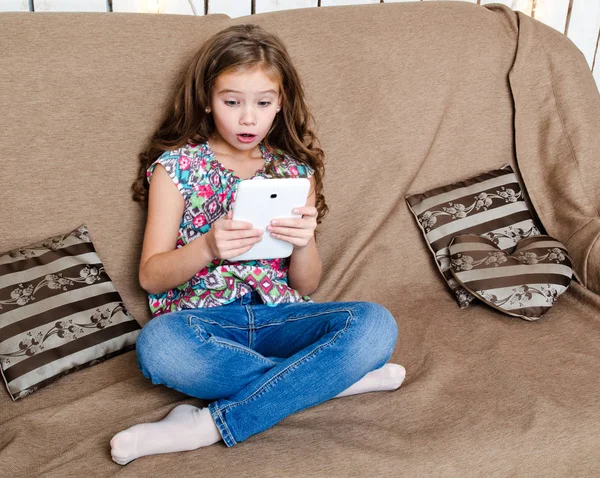 Симпатичная маленькая девочка с планшетным компьютером на диване дома — стоковое фото