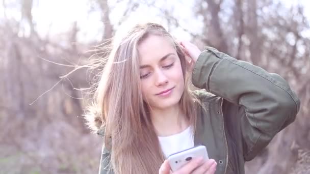 Retrato de mujer joven con el pelo rubio vestido con chaqueta sostiene un teléfono y está escribiendo un mensaje — Vídeos de Stock