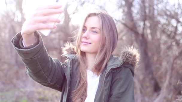 Joven chica hermosa con el pelo rubio vestido con una chaqueta y tomar una selfie en el parque y después de subir a un sitio web de medios sociales — Vídeo de stock