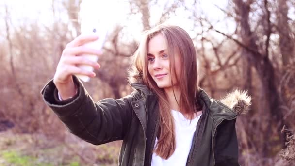 Menina europeia bonita com cabelo loiro vestido com uma jaqueta e tirar uma selfie no parque e depois de carregado para um site de mídia social — Vídeo de Stock