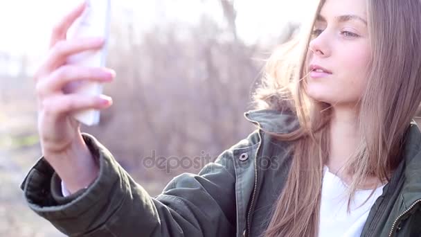 Hermosa mujer joven europea con el pelo rubio vestido con una chaqueta y tomar una selfie en el parque y después de subir a un sitio web de medios sociales — Vídeo de stock