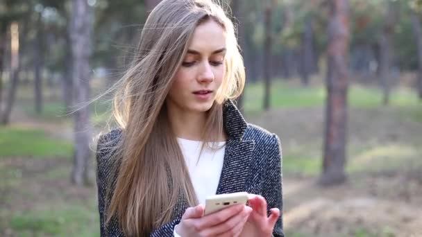 아름 다운 젊은 여자는 공원에 서 고 그녀의 휴대 전화에서 서핑 — 비디오