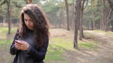 Kız telefonu cebinden çıkarır ve mesajı kontrol eder. Park açık havada mobil kullanarak uzun kıvırcık saçlı güzel esmer Kadın