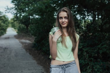 Young and beautiful girl with sporty body stands in park. She holds in hand cup with drinking straw.