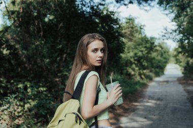 Young european woman looking over his shoulder at camera. Girl walks on parks road, on her shoulder has backpack. In hand woman holds a cup with drink.