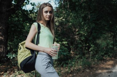 Avrupalı genç kadın parklar yolunda yürüyor. Kız el bardak tutar ve omzunda sarı bir sırt çantası var.