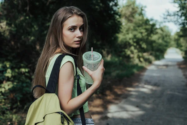 Young european woman looking over his shoulder at camera. Girl walks on parks road, on her shoulder has backpack. In hand woman holds a cup with drink.