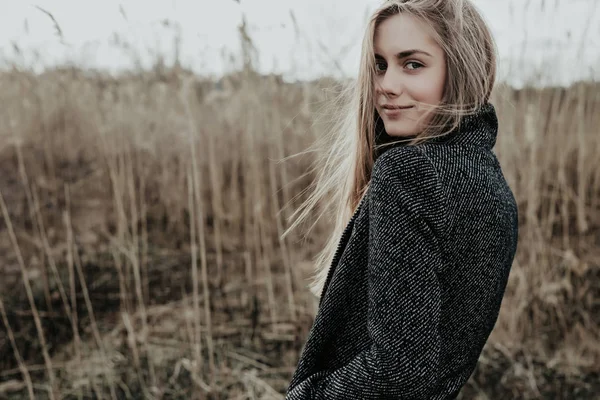 Uzun sarı saçlı hoş ve genç kadın onu kollamaya kameraya ve gülümseyen yün ceket giymiş. Arka plan bulrush. Sonbahar. Açık. Orta atış