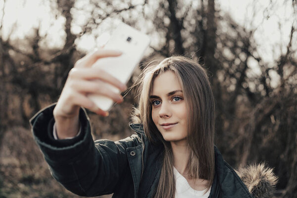 Pretty young woman taking selfie on mobile outdoor. Beautiful girl about twenty eyers old posing at camera in park. Model dressed in parka. Woman with blonde hair, full lips, wide eyebrows, blue eyes. Outdoor. Portrait.