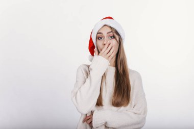 Noel Baba şapkası ağzını el ile kapsayan genç beyaz beyaz kadın. Kız bir şey tarafından şaşırttı. Genç ve güzel model sarı uzun saçlı, geniş kaşları ve mavi gözlü Studio fotoğraf makinesinde poz ile. izole.