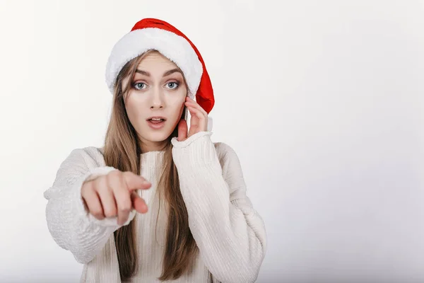 戴着圣诞老人帽子的女人用手指指着镜头 女孩惊讶的东西 漂亮的年轻模特 金发碧眼 宽眉和蓝眼睛在摄影棚里拍照 — 图库照片