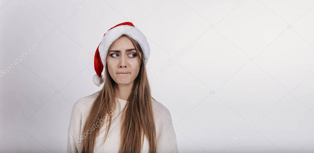  Woman in santa hat suspects something. Pretty young model with blonde long hair, wide eyebrows and blue eyes posing on camera in studio on white background. Isolated. Copy space. Place for advertising.