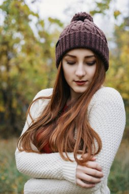 Yakışıklı genç kadınla kollarını birlikte mide üzerinde katlanmış. Sıcak kırmızı eşarp, kış şapka ve kazak modeli. Orada sonbahar park arka planda ayakta. Açık.