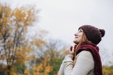 Sıcak kış şapka, kazak ve kırmızı eşarp ve gökyüzü üzerinde ararken model. Kız kollarını birlikte göğüs üzerinde katlanmış. Eşarp üzerinde ellerini tutan kadın. Açık. Profil portre.