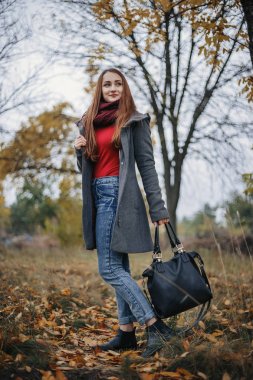 Mavi jeans, gri yün ceket, kırmızı kazak ve eşarp çanta sol elinde kamerayla poz güzel kadın. Kameraya bakarak modeli. Sonbahar. Islak hava. Açık