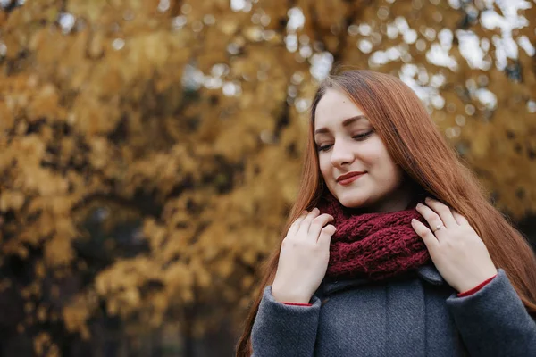 若いロマンチックな女性人は温かみのあるグレーのコートと赤いスカーフを身に着けています 背景が黄色の葉の木に秋の公園でモデル立ち 見ていると笑顔の少女です スカーフに彼女の手を保持している女性 — ストック写真
