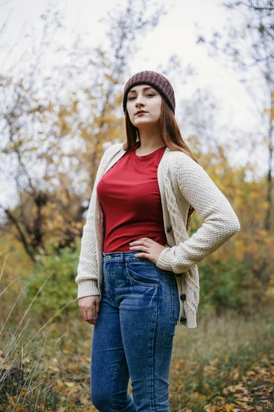 Young sexy woman with pretty face folded her arm on the hip and looking at camera. Model wearing warm red scarf, winter hat and sweater. She standing in autumn park background. Outdoor.