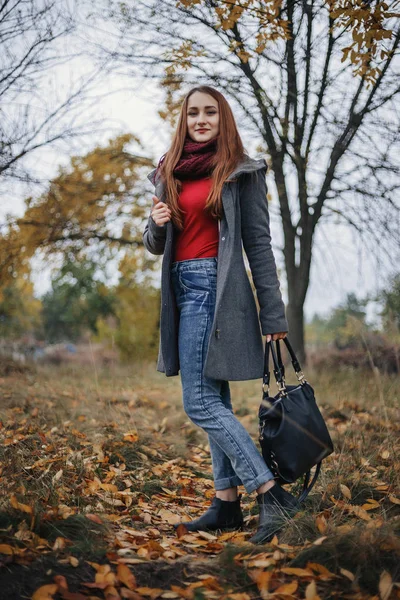 Pretty woman in blue jeans, grey wool coat, red sweater and scarf posing on the camera with bag in left hand. Model looking at camera. Autumn. Wet weather. Outdoor