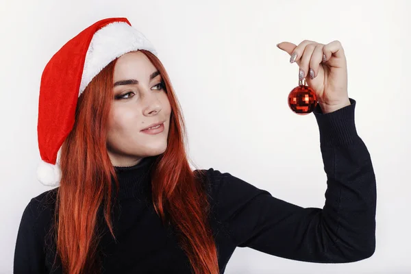 快乐的年轻红头发的女人在圣诞老人的帽子手拿着一个小圣诞球 并期待着它 — 图库照片