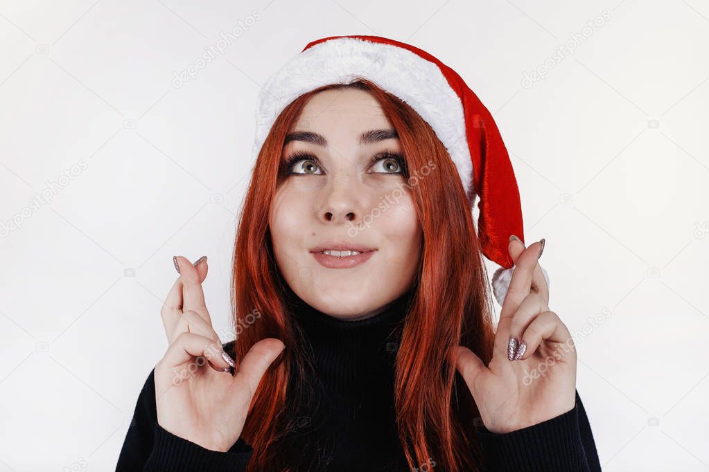 Happy young woman with readhead hair and in santa hat crossed her fingers 