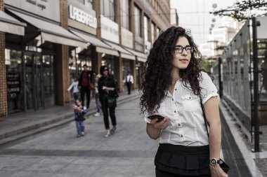 Şehir arka plan üzerinde duran genç karışık ırk kadın. Sokakta duran ve yan bakarak kadın. Cep telefonu elinde tutan genç bayan